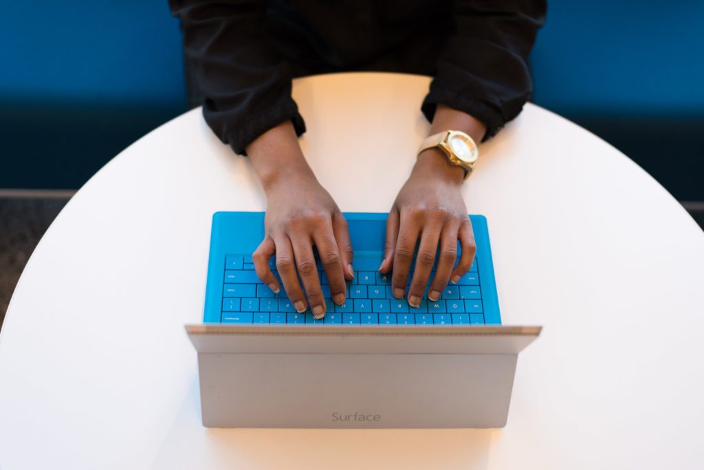 Woman wearing gold watch, sitting at table, typing on a Microsoft Surface notebook