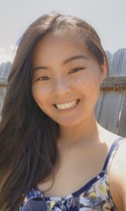Color photograph of Emily Ehlen smiling in the sunshine with her long dark hair covering one shoulder while wearing a floral-printed top